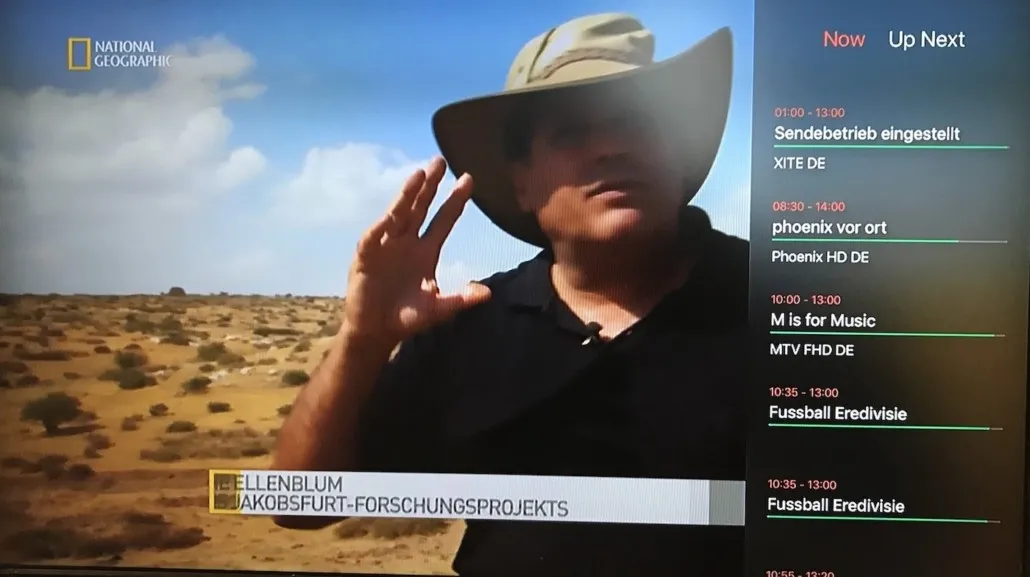 Vista de canal en directo de iPlayTV en Apple TV con la superposición de la guía de programación Ahora y A continuación
